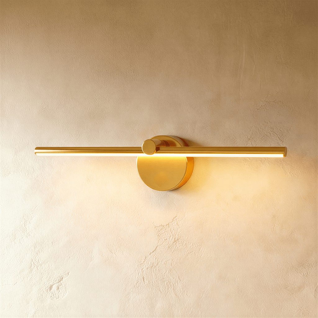 Clérachic wall lamp in brushed gold, its slim bar glowing warm white above a round switch-knob backplate.
