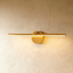Clérachic wall lamp in brushed gold, its slim bar glowing warm white above a round switch-knob backplate.