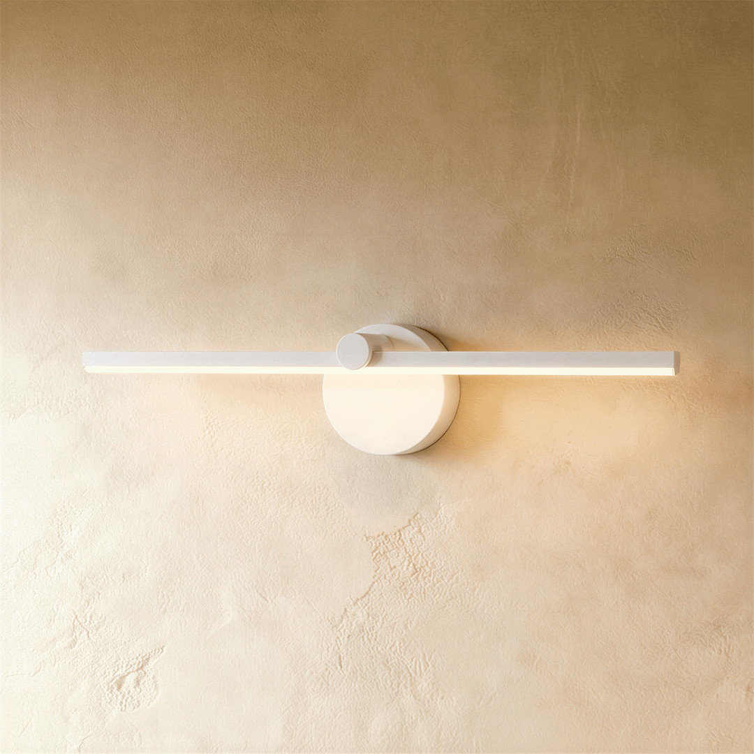 Clérachic wall lamp in white, its slender bar glowing warm white beside a round switch-knob wall plate.