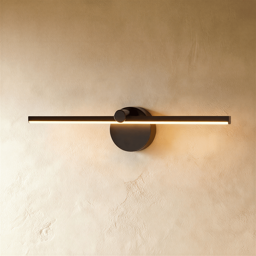 Clérachic wall lamp in matte black, the slim linear bar glowing warm white against a plaster wall.