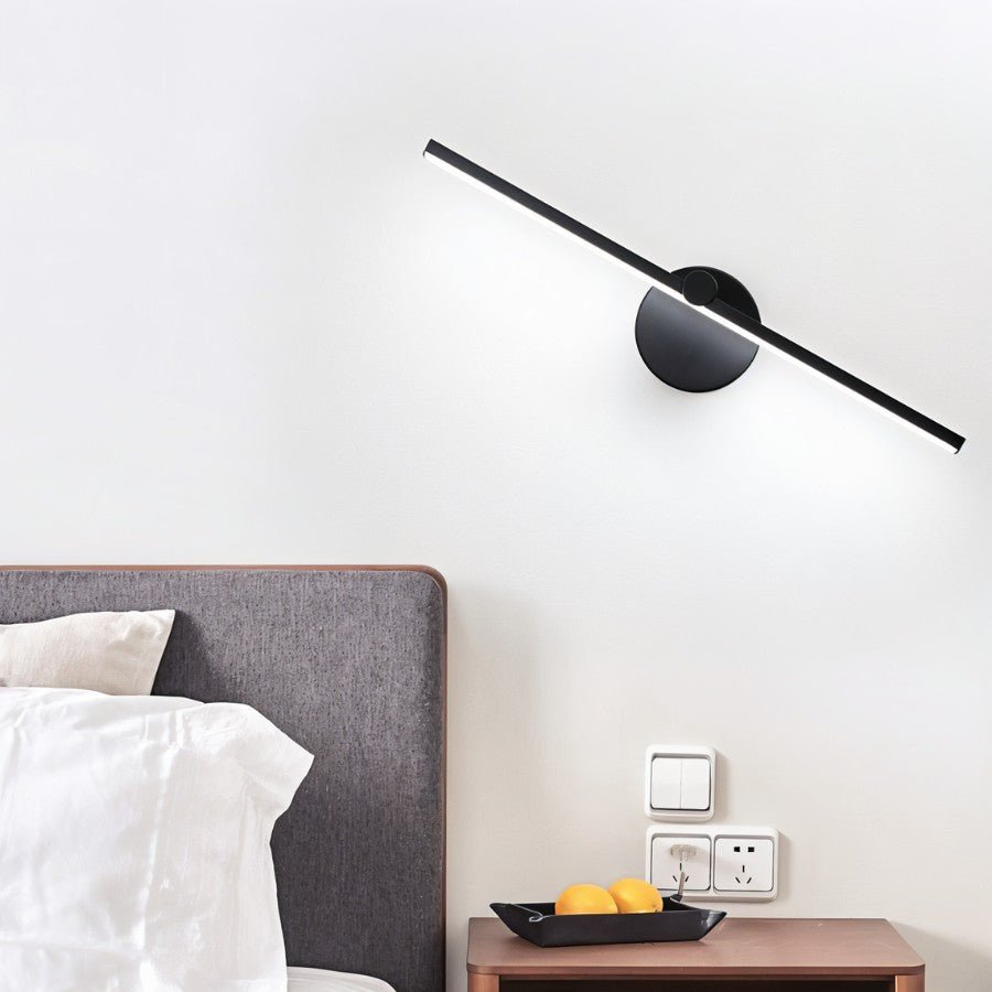 Clérachic wall lamp in black mounted at an angle above a bedside table, its bar glowing warm white in a bedroom.
