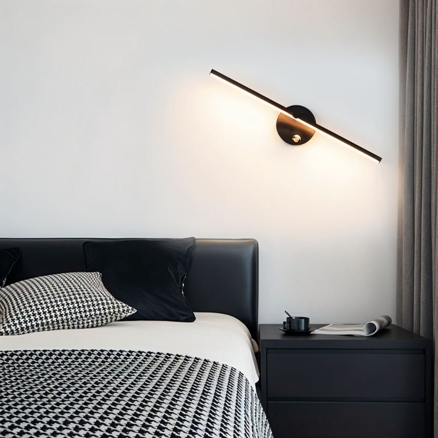 Clérachic wall lamp in black tilted above a dark bedside table, its bar glowing warm white over houndstooth bedding.