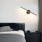 Clérachic wall lamp in black tilted above a dark bedside table, its bar glowing warm white over houndstooth bedding.