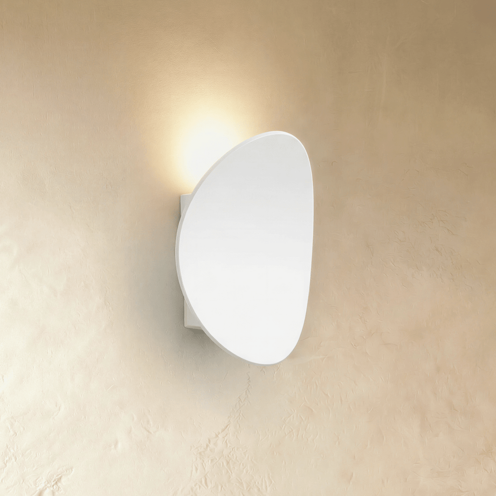 Clérachic's white pebble-shaped wall light throws soft warm light up a plaster wall in profile view.