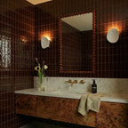 Two Clérachic white wall lights flank a bronze-tiled bathroom mirror above a marble vanity and dark wood cabinet.