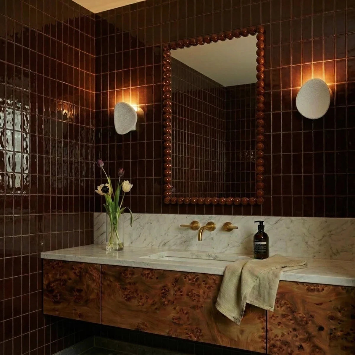 Two Clérachic white wall lights flank a bronze-tiled bathroom mirror above a marble vanity and dark wood cabinet.