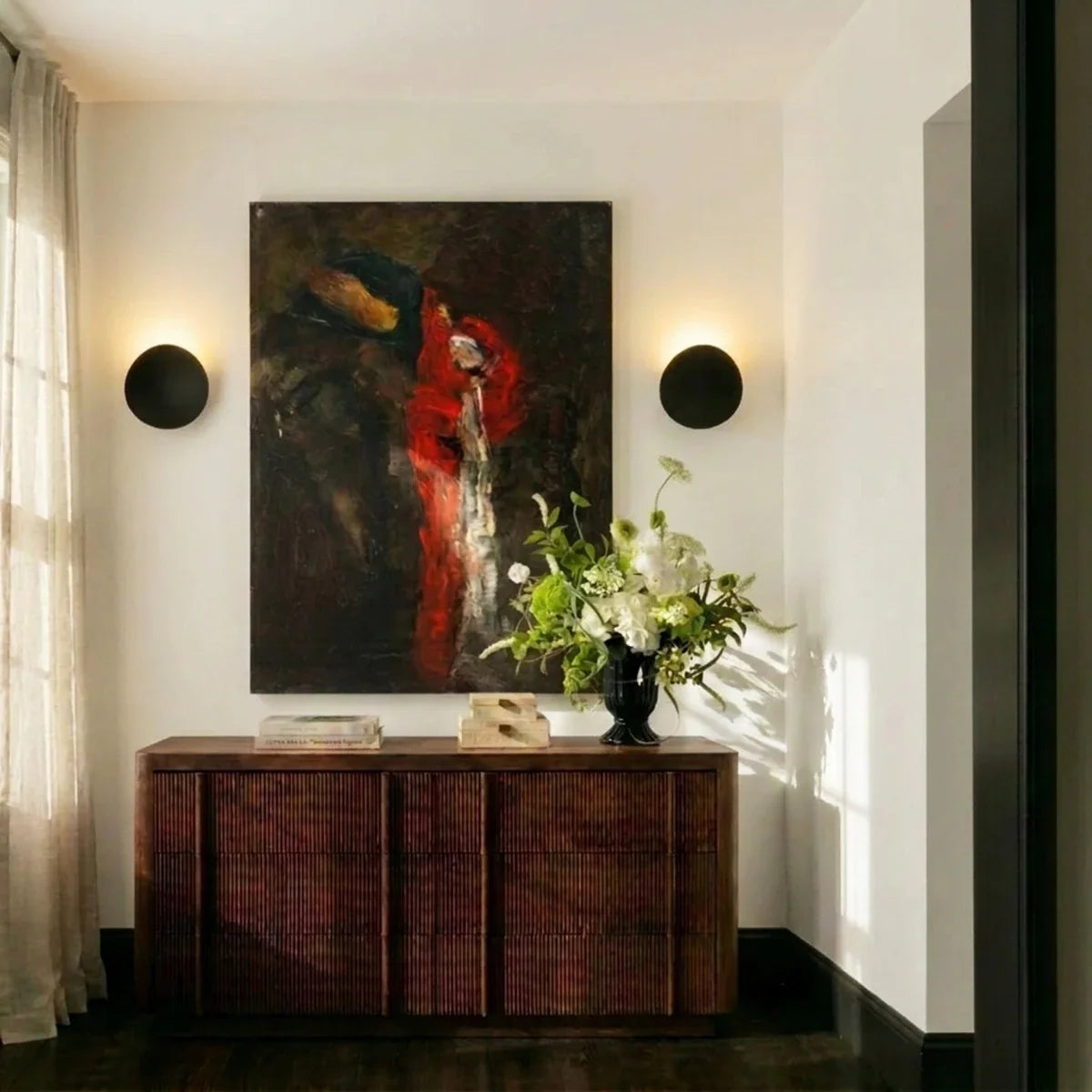Two Clérachic black wall lights flank a large abstract painting above a dark wood sideboard with fresh flowers.