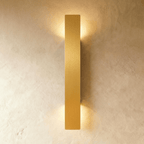 Clérachic brushed gold aluminium wall light, slim bar profile, glowing warm against a textured plaster wall.
