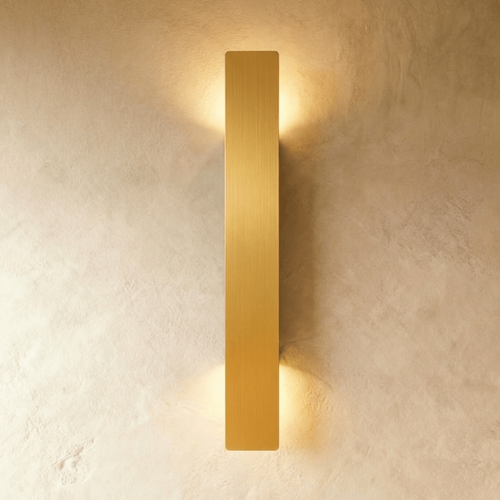 Clérachic brushed gold aluminium wall light, slim bar profile, glowing warm against a textured plaster wall.