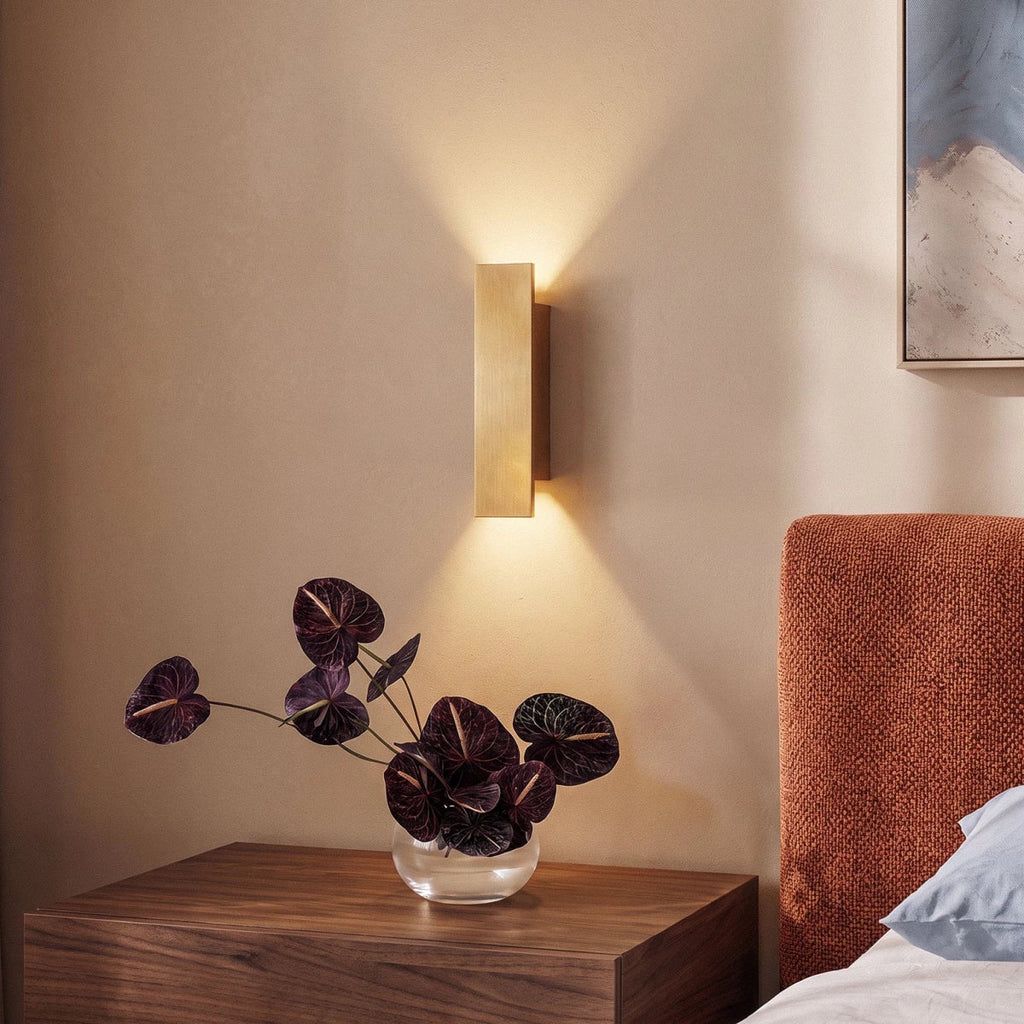 Clérachic brushed gold wall light mounted above a walnut nightstand, warm light framing dark anthurium leaves.