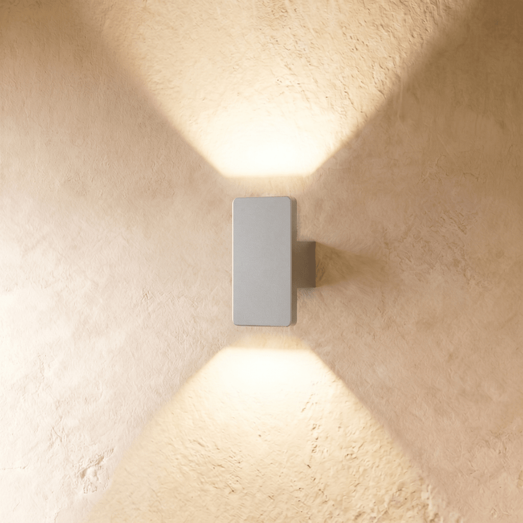 A compact Clérachic white wall light casts warm up-and-down beams across a sunlit plastered wall surface.