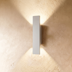 A taller Clérachic white wall light throws warm up-and-down beams across a textured plastered wall surface.