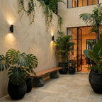 Two Clérachic black wall lights glow at dusk along a stone courtyard wall lined with potted palms and a bench.