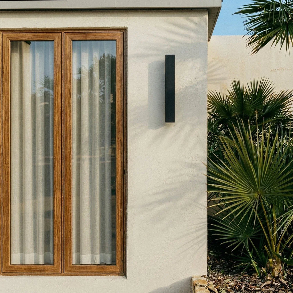 A Clérachic black wall light sits beside a timber-framed window, palm fronds catching afternoon light nearby.