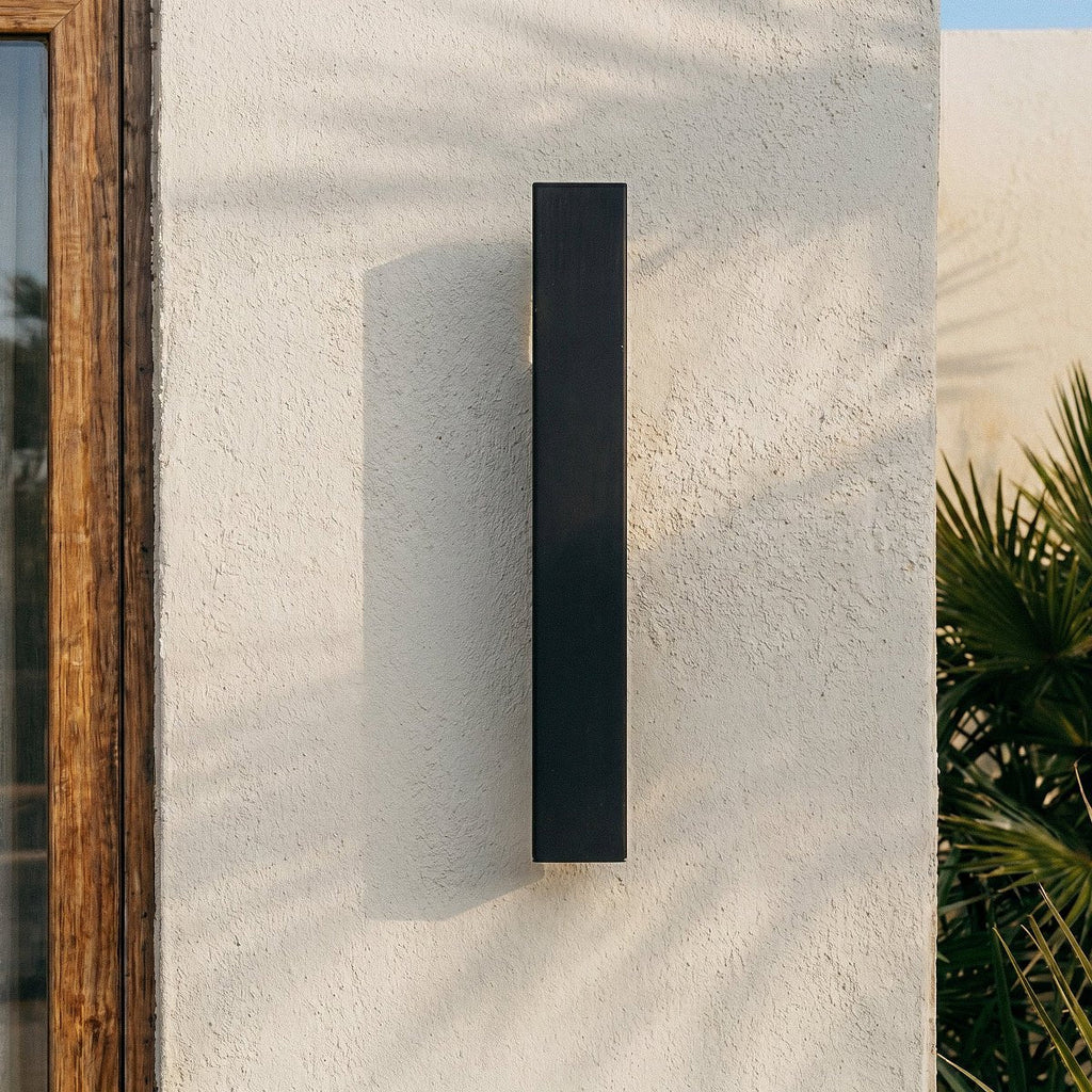 Close-up on the Clérachic black wall light mounted to a stucco corner beside a wood door and palm fronds.