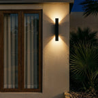 At night, the Clérachic black wall light glows warmly beside a curtained window and dark green palm leaves.