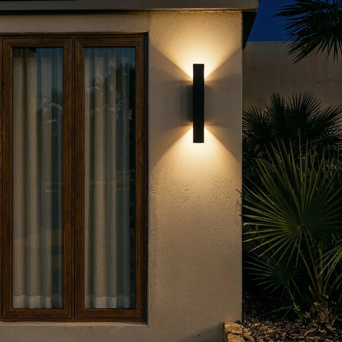 At night, the Clérachic black wall light glows warmly beside a curtained window and dark green palm leaves.