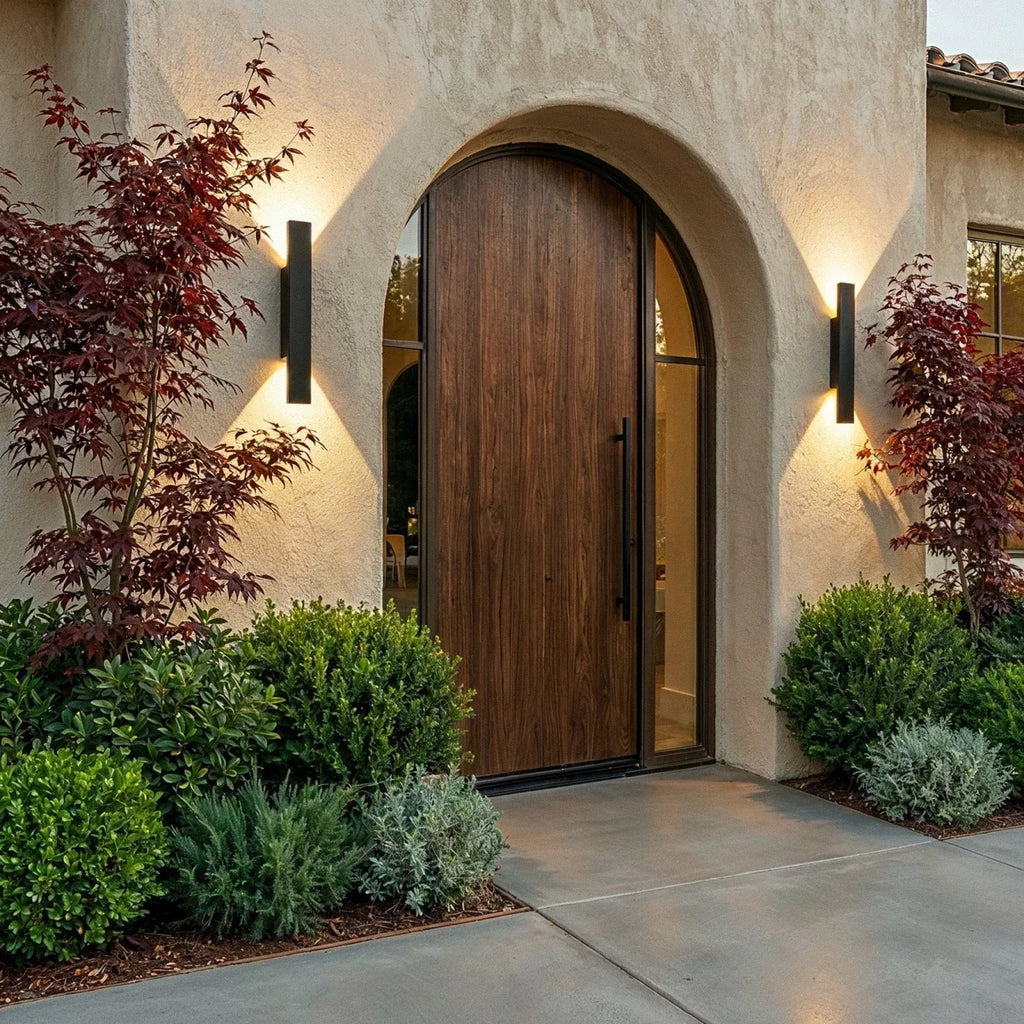 Two Clérachic black wall lights flank an arched wood door at dusk, framed by red maple leaves and boxwood.
