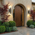 Two Clérachic black wall lights flank an arched wood door at dusk, framed by red maple leaves and boxwood.