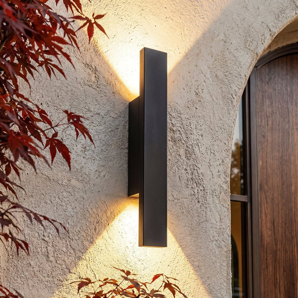 Close-up on the Clérachic black wall light beside the arched wood door, warm light catching red maple leaves.