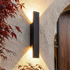 Close-up on the Clérachic black wall light beside the arched wood door, warm light catching red maple leaves.