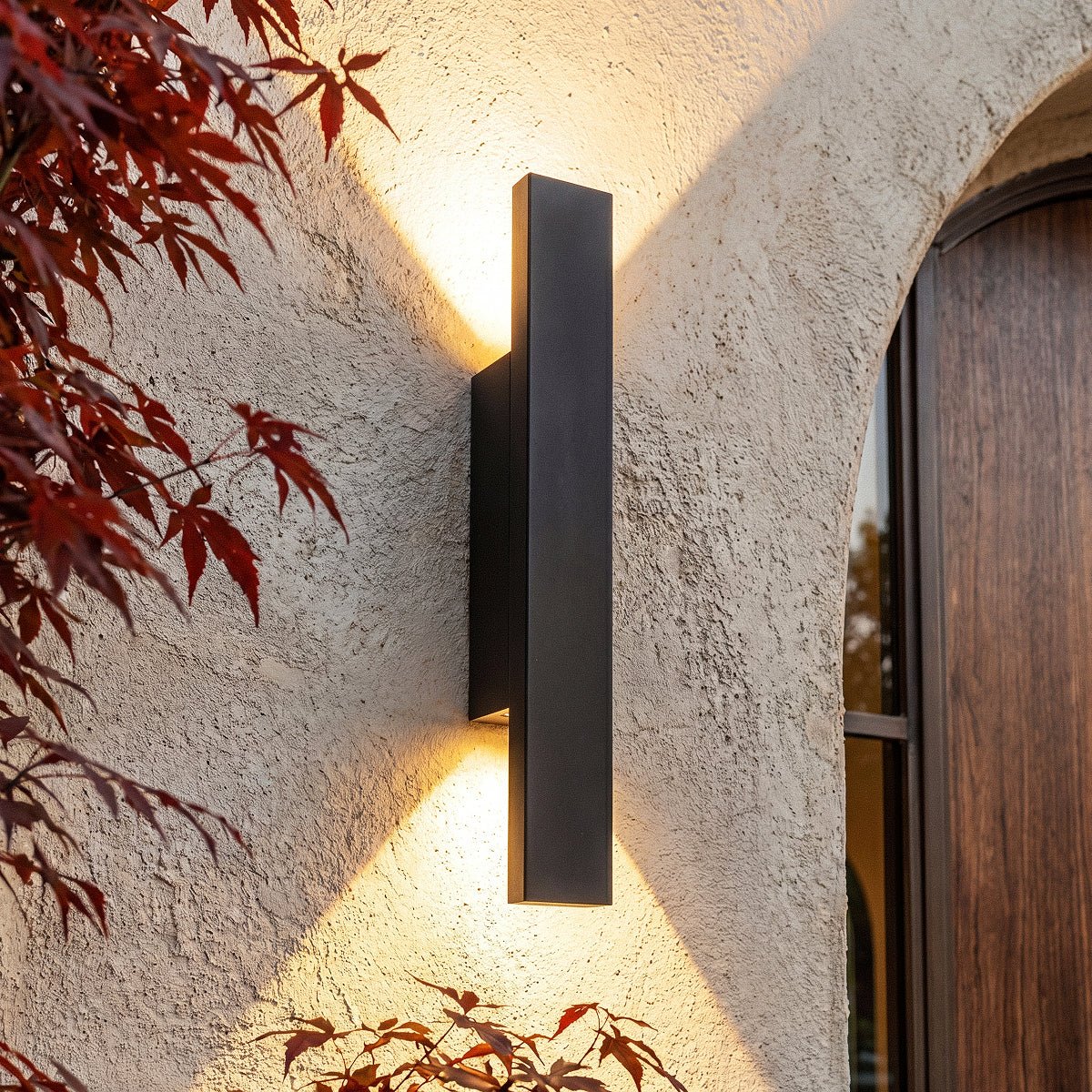 Close-up on the Clérachic black wall light beside the arched wood door, warm light catching red maple leaves.