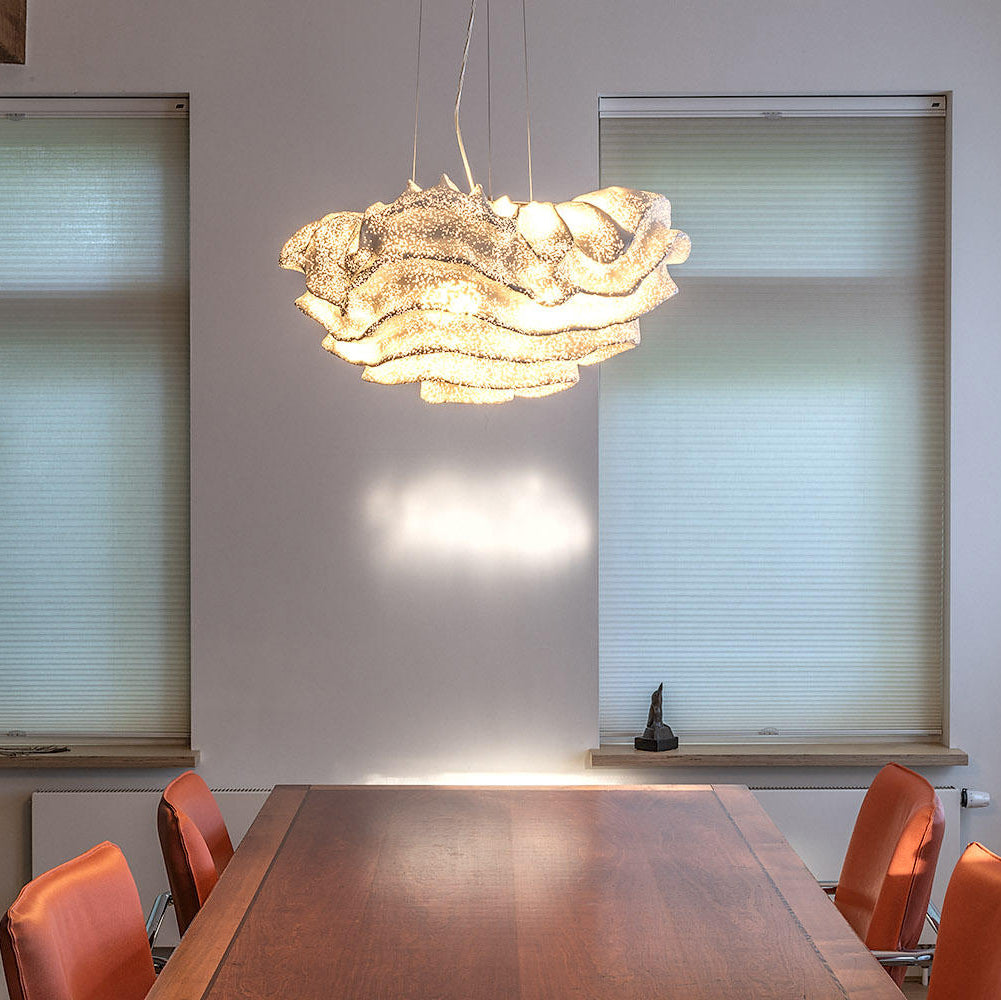 Lit warm, a Clérachic ruffled pendant hangs above a polished wooden dining table with orange chairs, beside a pale blinded window.