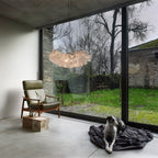 A Clérachic pendant with a ruffled white shade hangs by a floor-to-ceiling window, near an olive armchair and a dog resting on a dark blanket.
