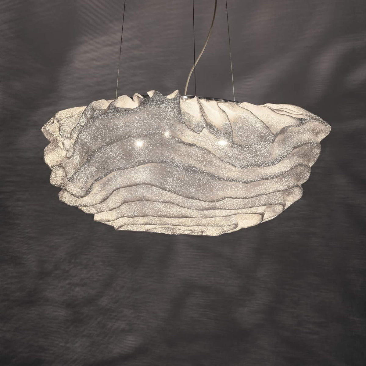 The ruffled white shade of this Clérachic pendant glowing against a dark grey wall marked with soft shadow patterns.
