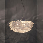 Warm light glows through the layered shade of this Clérachic pendant, set against a dark grey wall with soft ripples of shadow.