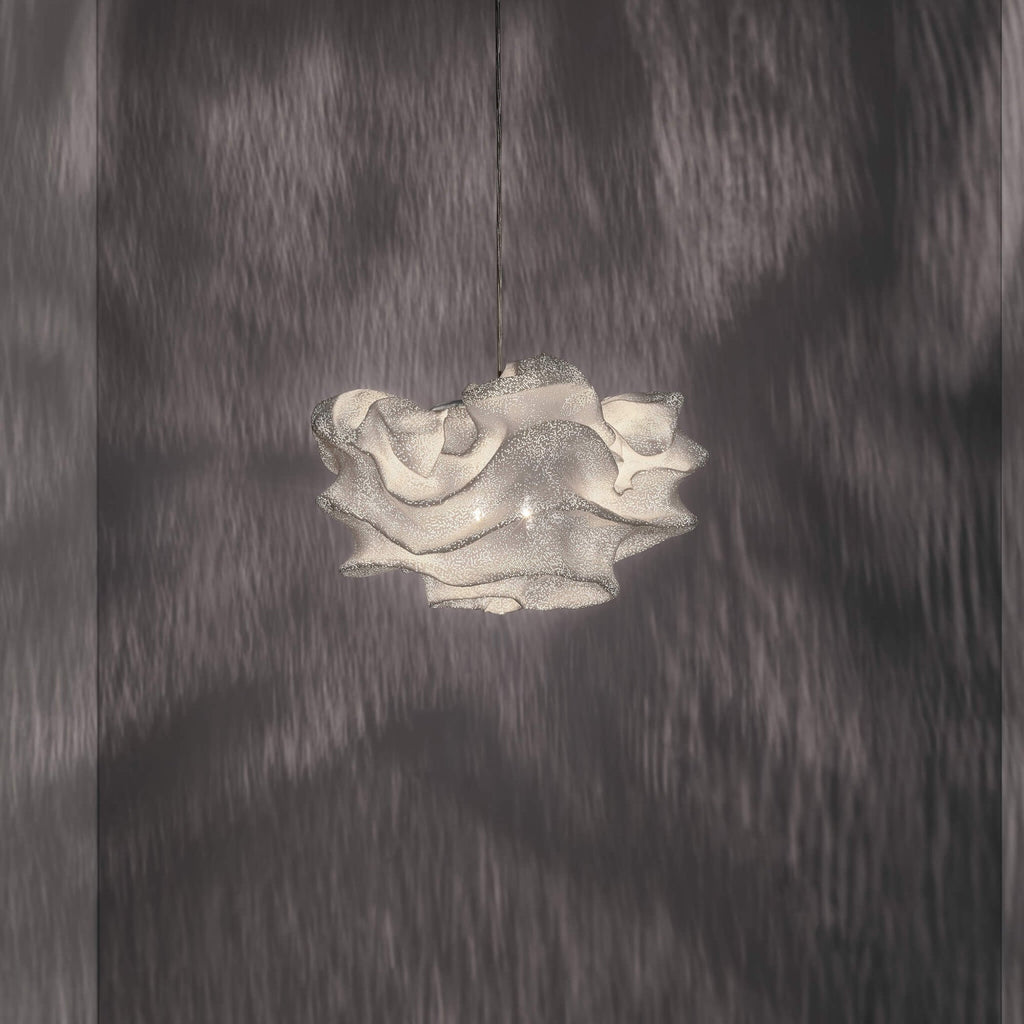 A Clérachic pendant with a crinkled white mesh shade, lit and hanging on a dark wall of soft shadow patterns.