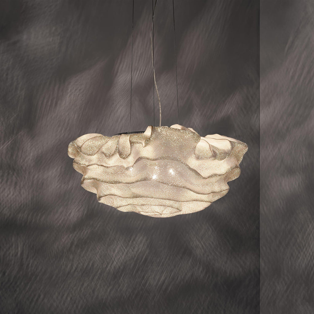 This Clérachic pendant glowing warm, its layered shade lit from inside, set against a dark grey wall.