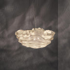 This Clérachic pendant glowing warm, its layered shade lit from inside, set against a dark grey wall.