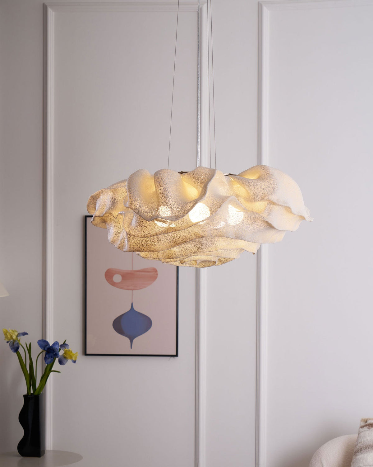 Warm light from a Clérachic pendant with an ivory ruffled shade, hung in a white panelled room with a framed print and fresh irises.