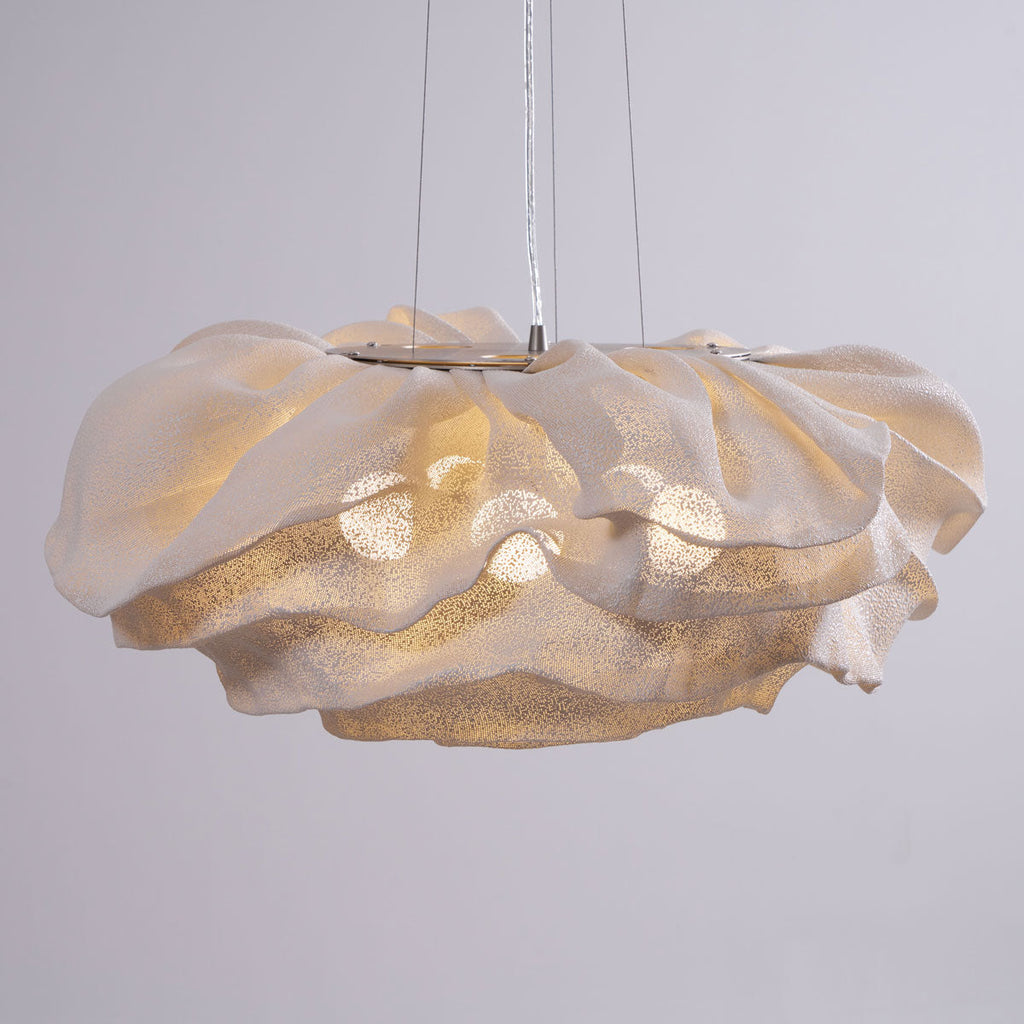 A Clérachic pendant with a wide ruffled shade, lit warm, hanging on thin wires against a pale lilac-grey wall.