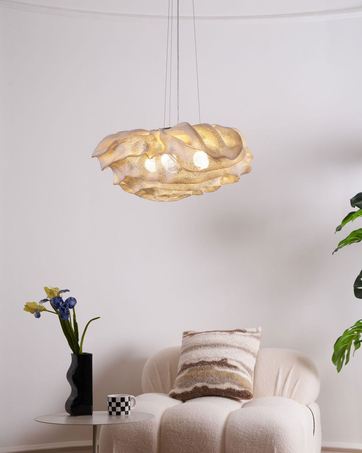High above a cream bouclé armchair, a Clérachic pendant glows warm, with a vase of irises and a plant beside it.