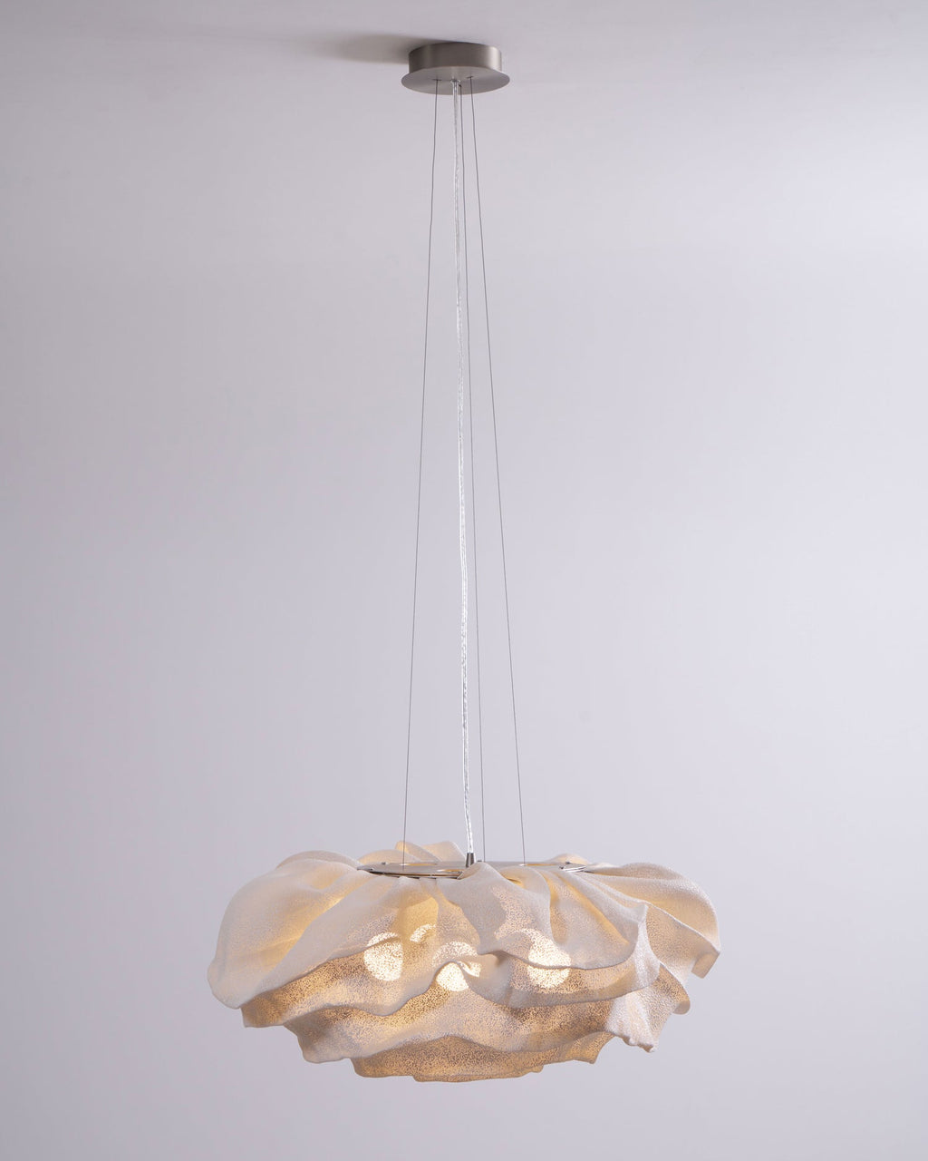 A Clérachic pendant hangs from a round ceiling plate on fine wires, its ruffled shade glowing against a pale grey wall.
