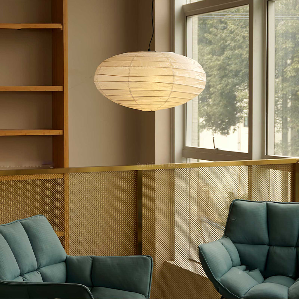 Clérachic oval paper pendant light lit beside teal armchairs and a gold mesh balustrade, a window with trees beyond.
