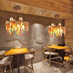 Two Clérachic wine bottle chandeliers hung in a rustic dining space, their round frames lit above wood tables against a textured wall.