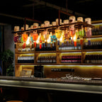 The Clérachic Wine Bottle Glass Pendant Light glows over a bar, rust-toned frame packed with colored bottles.