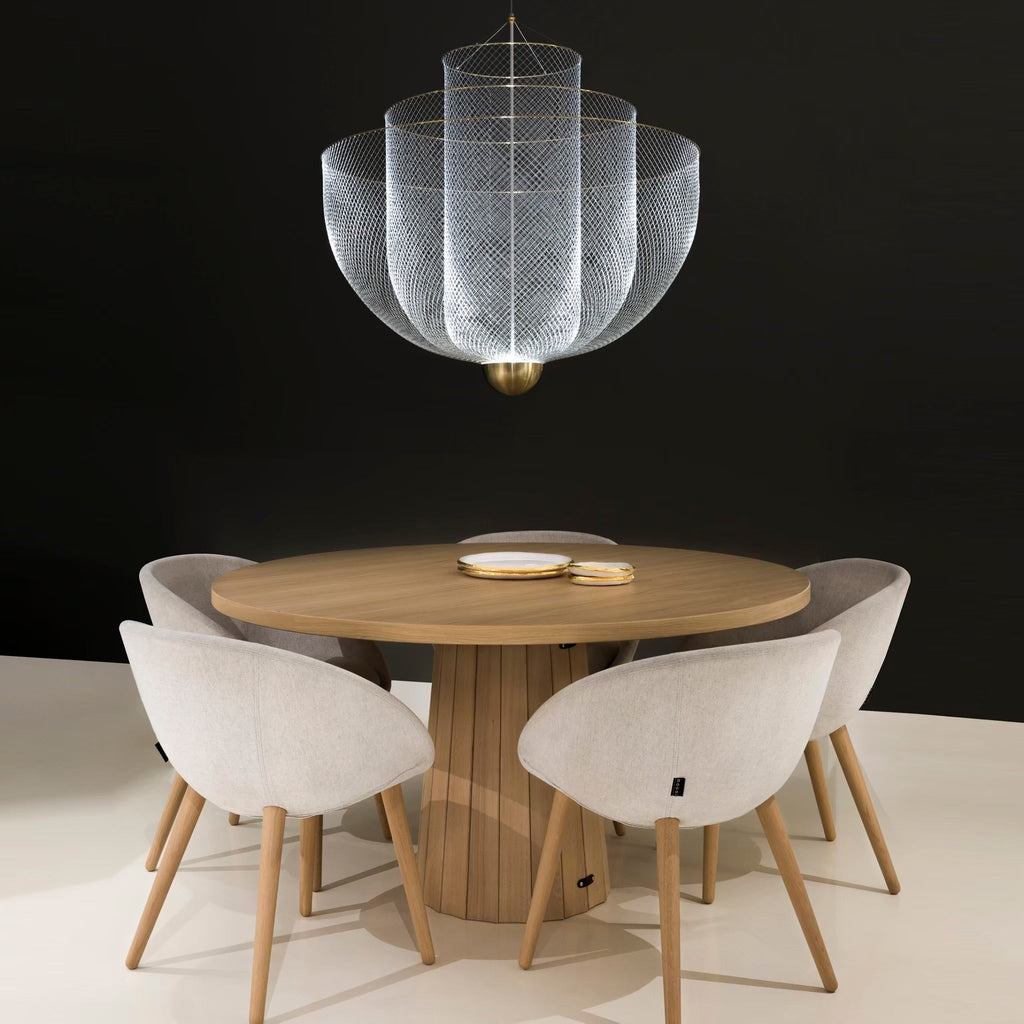 A silver wire-mesh Clérachic chandelier above a round wooden table with pale upholstered chairs, against a black wall.