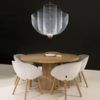 A silver wire-mesh Clérachic chandelier above a round wooden table with pale upholstered chairs, against a black wall.