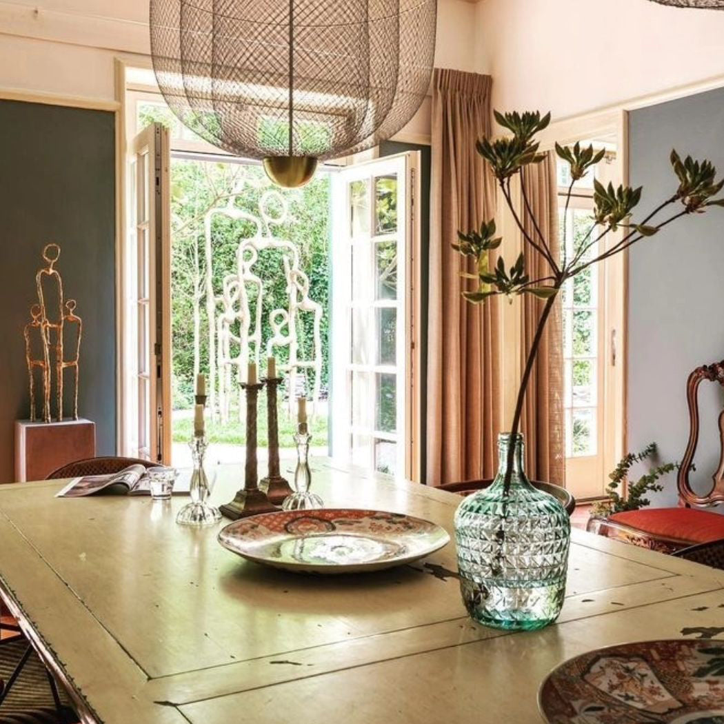 A Clérachic mesh chandelier above a table with a green glass vase and a plate, in front of open doors and an apricot curtain.