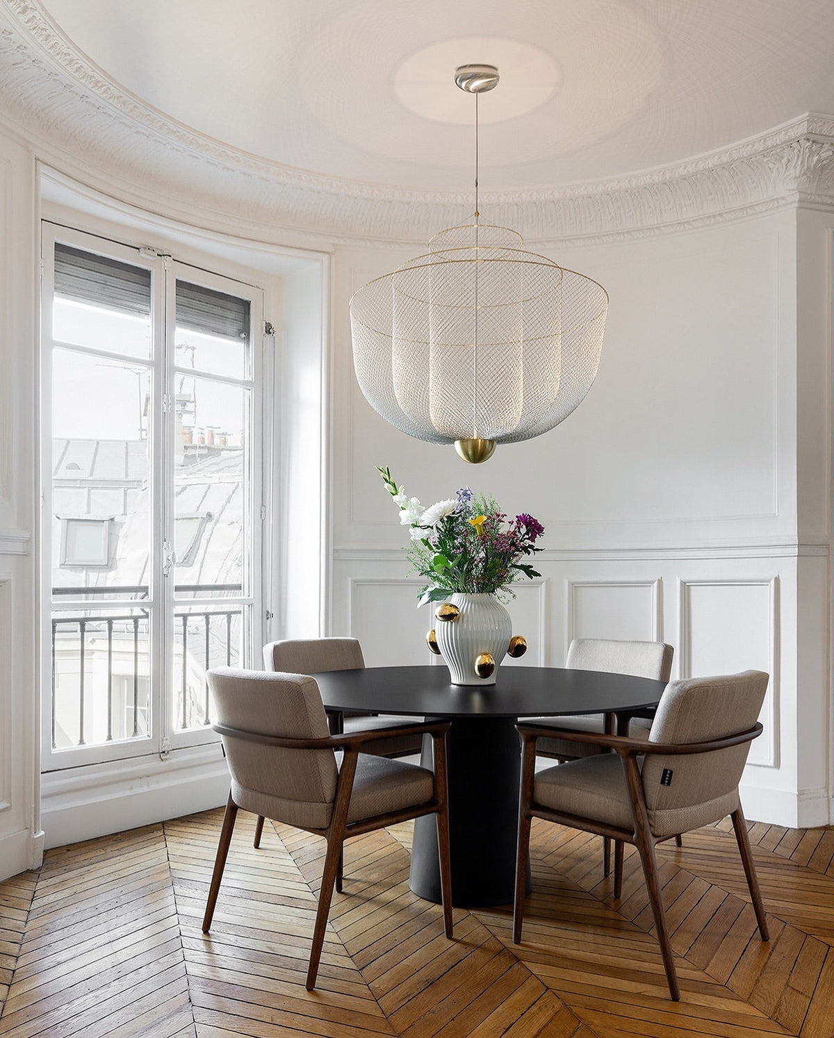 Clérachic wire-mesh chandelier in a white panelled room with tall windows, above a dark round table with a bouquet of flowers.
