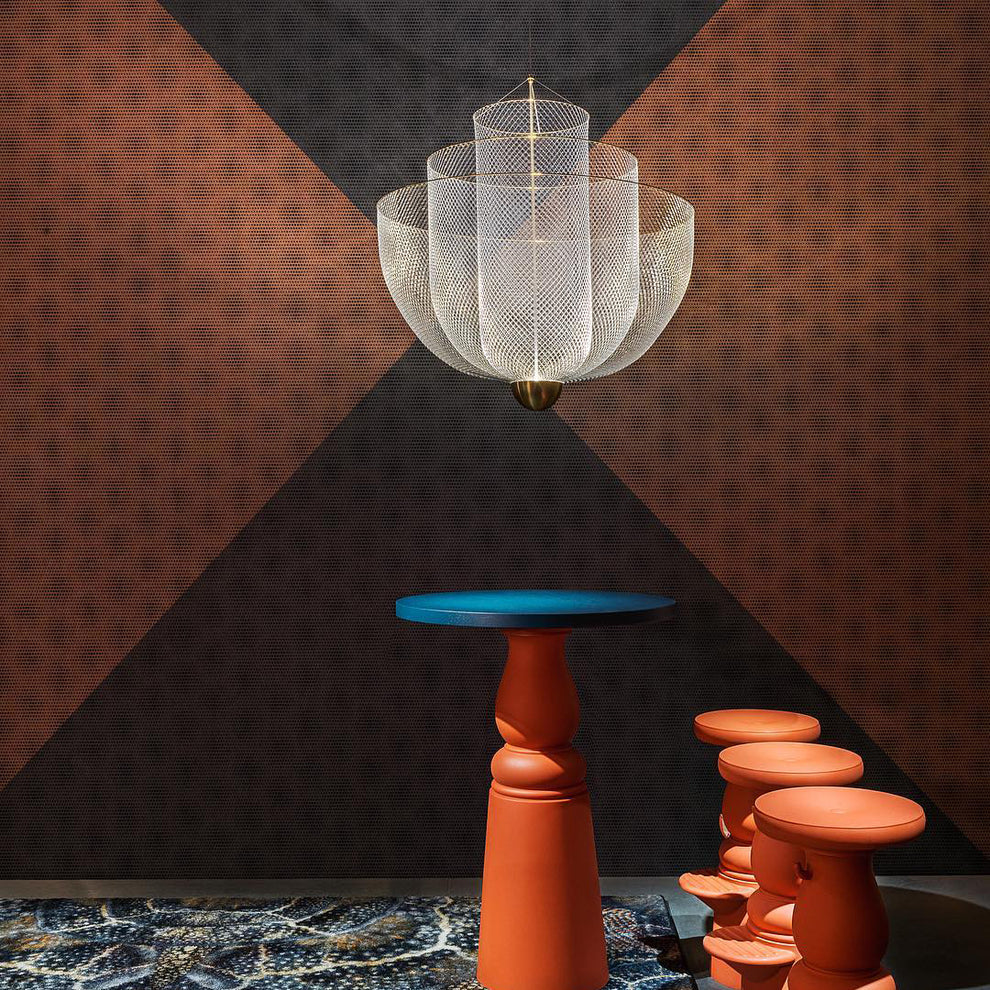 A Clérachic mesh chandelier, unlit, hung on a rust and charcoal wall, with a blue-topped pedestal and orange stools beneath.