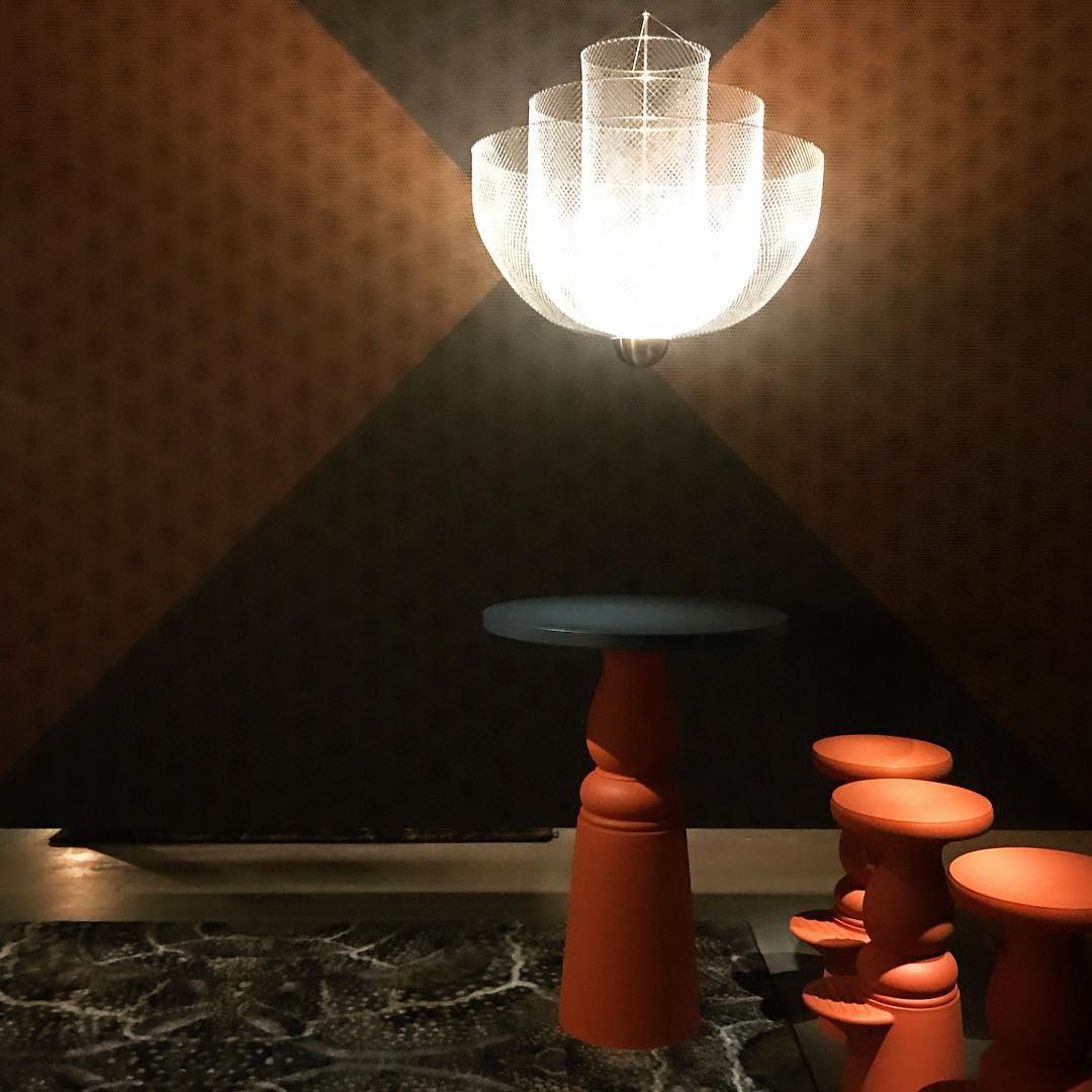 The same Clérachic mesh chandelier lit with a bright white glow, against a rust and charcoal wall in a dim room.