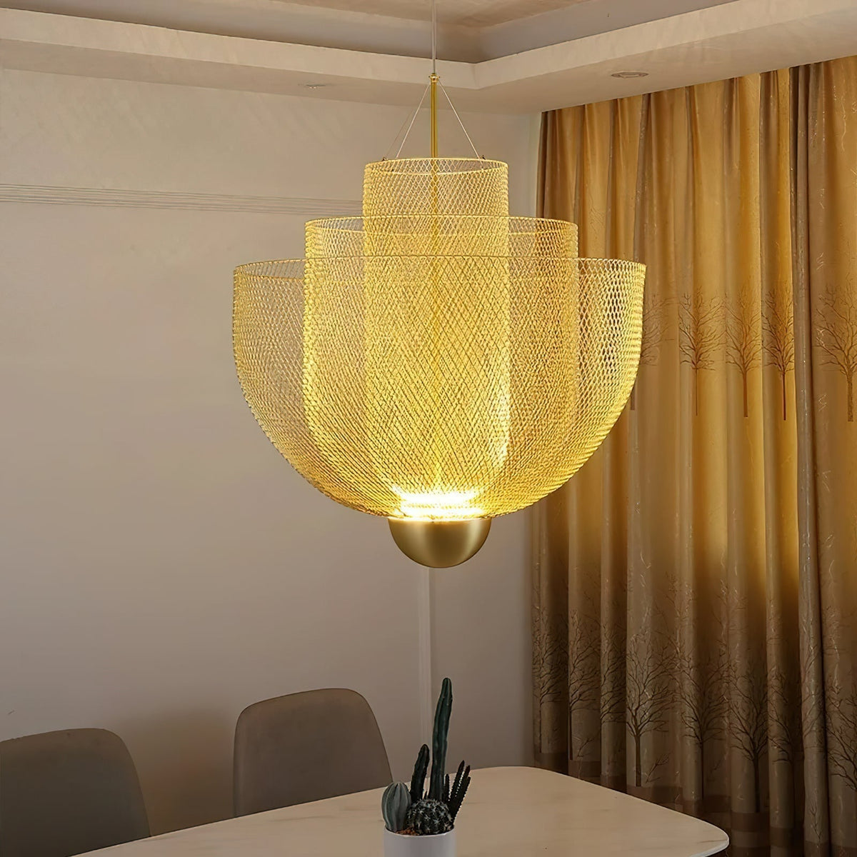 Clérachic gold mesh chandelier glowing from within, hung in front of a cream wall and gold curtains, with a cactus below.