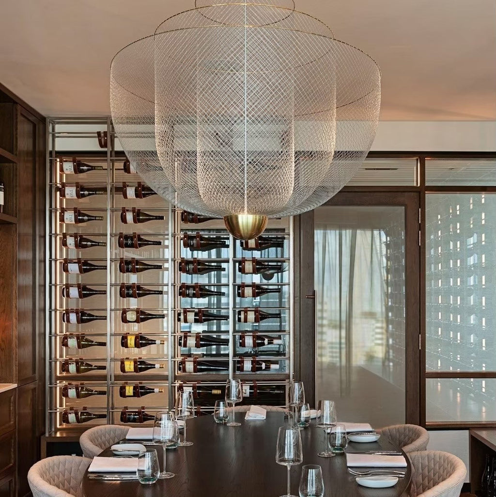 Clérachic wire-mesh chandelier above a long dark table set with glasses, in front of a wall of wine bottles.