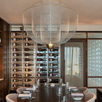 Clérachic wire-mesh chandelier above a long dark table set with glasses, in front of a wall of wine bottles.
