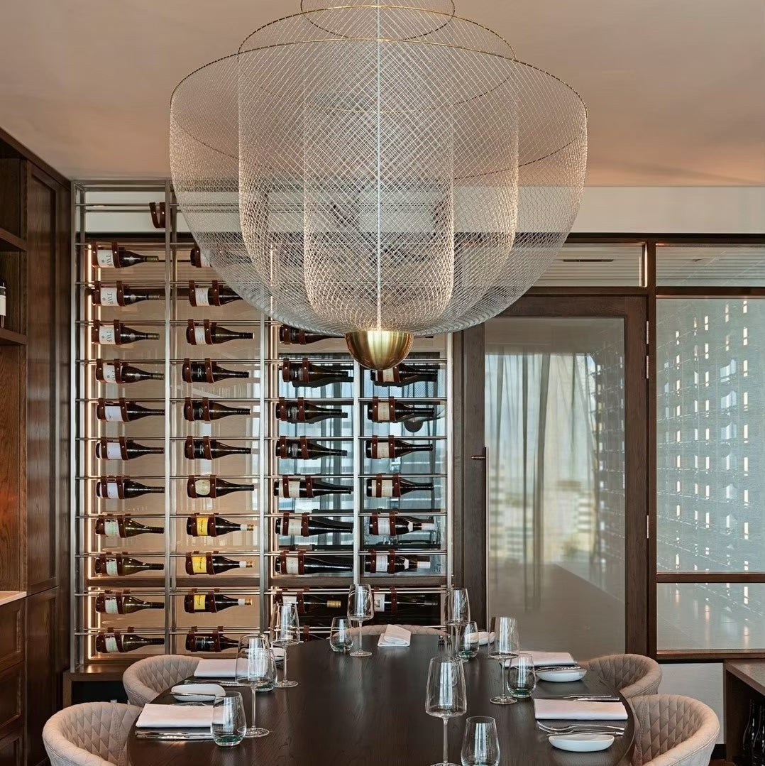 Clérachic wire-mesh chandelier above a long dark table set with glasses, in front of a wall of wine bottles.